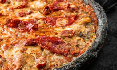 Siyah hamurlu pizza Pepperoni, jambon, sosis, parmesan peyniri, taştan siyah arka planda ıspanak. Piknik şirketi için fast food, üst görünüm, seçici odaklanma.