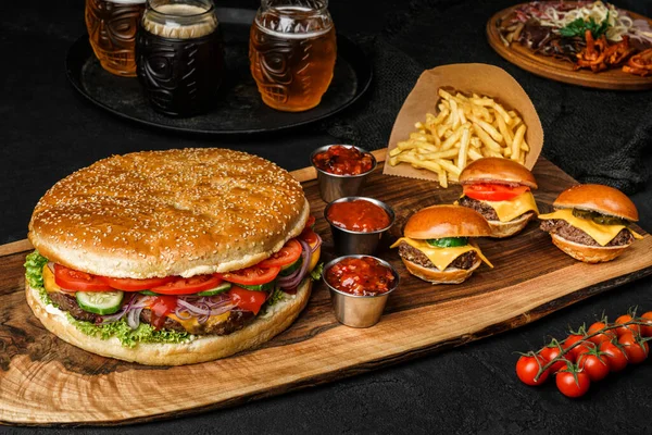 Big burger and classic burgers with meat cutlets, cheese, tomatoes, cucumbers, lettuce and sauce on wooden board with french fries potatoes and beer on black background. Fast food for picnic