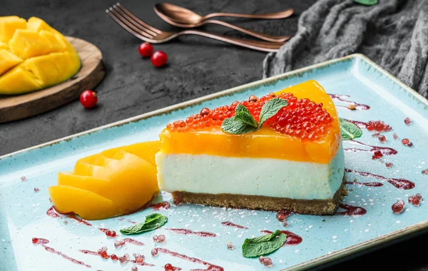 Creamy mango cheesecake decorated with red berries and mint on a large plate and dark background, close up view. Summer desserts and sweets