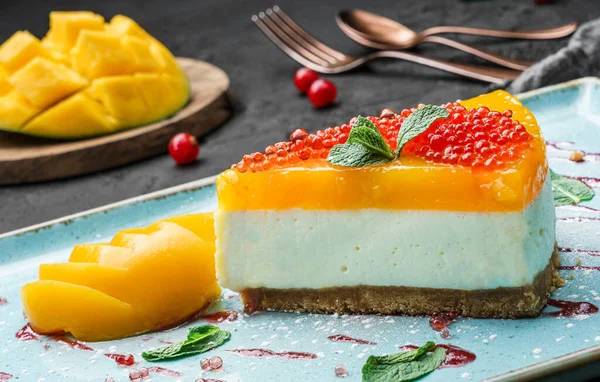 Creamy mango cheesecake decorated with red berries and mint on a large plate and dark background, close up view. Summer desserts and sweets