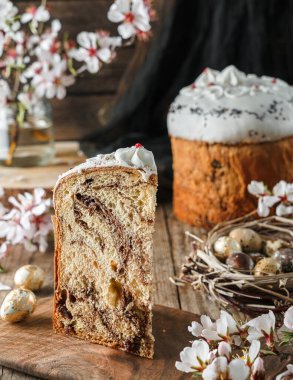 Slice of Homemade glazed Easter cake or bread kulich decorated with candy on rustic wooden table with easter eggs in nest and spring flowers. Happy Easter holiday