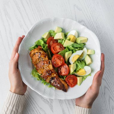 Ellerinde mangalda tavuk göğsü dilimleri, domates, yumurta, avokado, marul ve hafif ahşap zemin üzerinde tohum olan Buda salatası var. Sağlıklı yemek, temiz yemek, diyet, üst görüş.