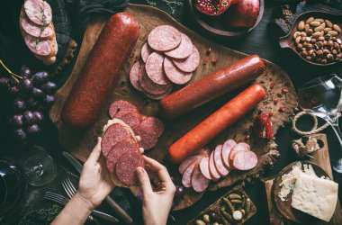 Koyu arkaplanda tahta bir kesme tahtasının üzerinde salam dilimli sandviçi tutan eller. Sosisli, jambonlu, salamlı, peynirli, meyveli, şaraplı, fındıklı meze. Üst görünüm, tonlama