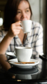 Zblízka záběr Bílý hrnek na kávu je umístěn na stole, mladá žena sedí pít kávu v pozadí.