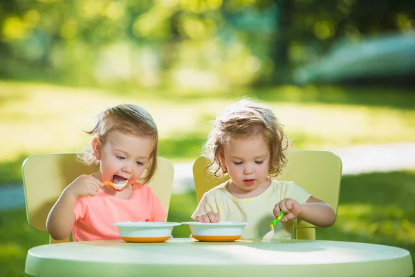 Two little girls sitting at a table and eating together against green lawn — Stock Photo, Image