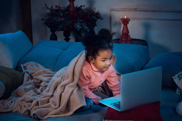 Happy african-american little girl during video call with laptop and home devices, looks delighted and happy