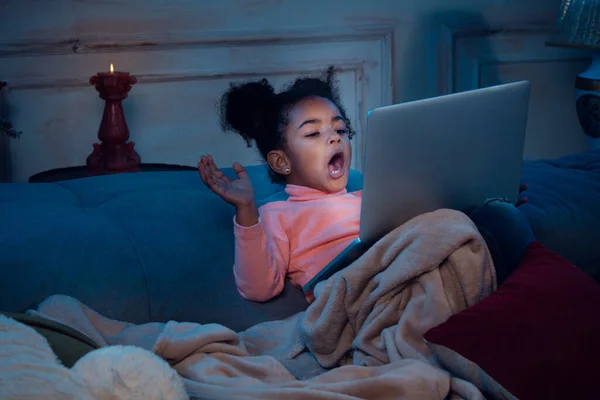 Happy african-american little girl during video call with laptop and home devices, looks delighted and happy