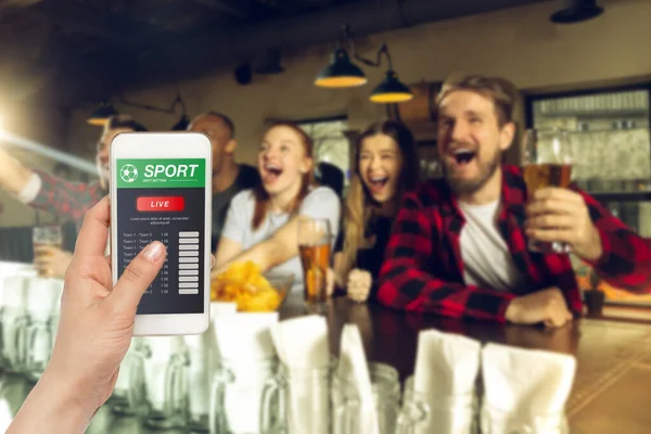 Device screen with mobile app for betting and score, cheering friends, fans on background