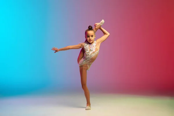 Mała kaukaska dziewczyna, gimnastyka rytmiczna trening, wykonywania izolowane na gradientowym tle niebiesko-czerwony studio w neonie — Zdjęcie stockowe