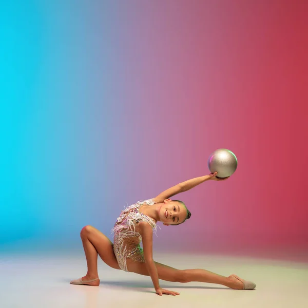 Little caucasian girl, rhytmic gymnast training, performing isolated on gradient blue-red studio background in neon — Stock Photo, Image