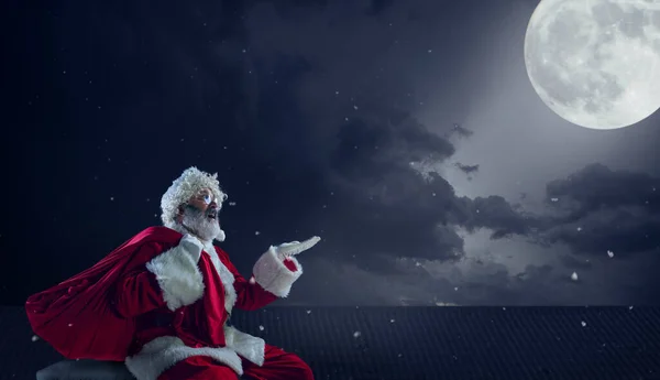 Emotional Santa Claus congratulating with New Year and Christmas, sitting on roof of the house in midnight with full moon