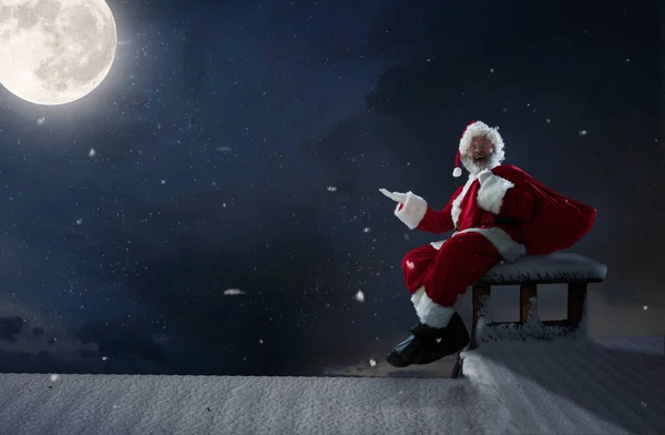 Emocjonalny Święty Mikołaj gratuluje Nowego Roku i Bożego Narodzenia, siedzi na dachu domu w nocy z pełnią księżyca — Zdjęcie stockowe