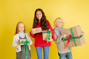 Noel tatilinde hediye vermek ve almak. Mutlu gülümseyen bir grup çocuk sarı stüdyo arka planında izole edilmiş eğleniyor.
