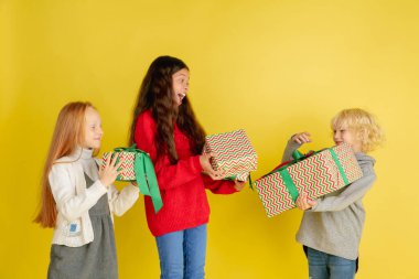 Noel tatilinde hediye vermek ve almak. Mutlu gülümseyen bir grup çocuk sarı stüdyo arka planında izole edilmiş eğleniyor.