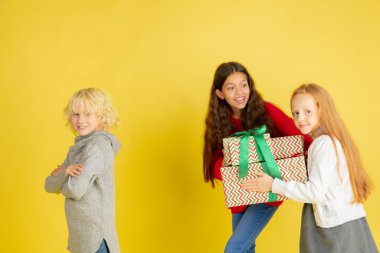 Noel tatilinde hediye vermek ve almak. Mutlu gülümseyen bir grup çocuk sarı stüdyo arka planında izole edilmiş eğleniyor.