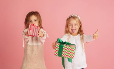 Noel tatilinde hediye vermek ve almak. İki küçük gülümseyen çocuk pembe stüdyo arka planında izole edilmiş eğleniyor.