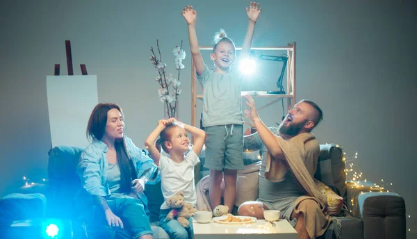 Šťastná rodina sleduje projektor, televizi, filmy s popcornem ve večerních hodinách doma. Matka, otec a děti spolu tráví čas. — Stock fotografie