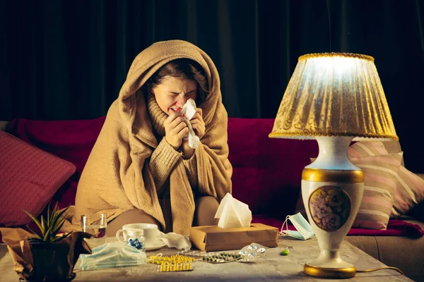 Woman wrapped in a plaid looks sick, ill, sneezing and coughing sitting at home indoors