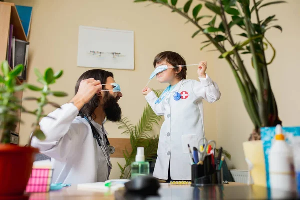 Médico pediatra examinando a un niño mientras usa mascarilla en un cómodo consultorio médico — Foto de Stock