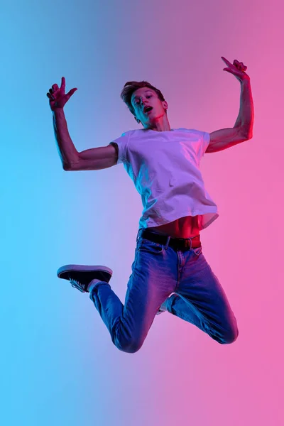 Young caucasian mans jumping high on gradient blue-pink studio background in neon light