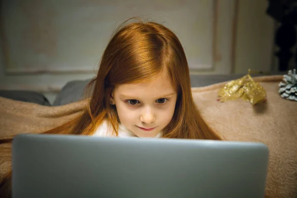 Happy caucasian little girl during video call or messaging with Santa using laptop and home devices, looks delighted and happy