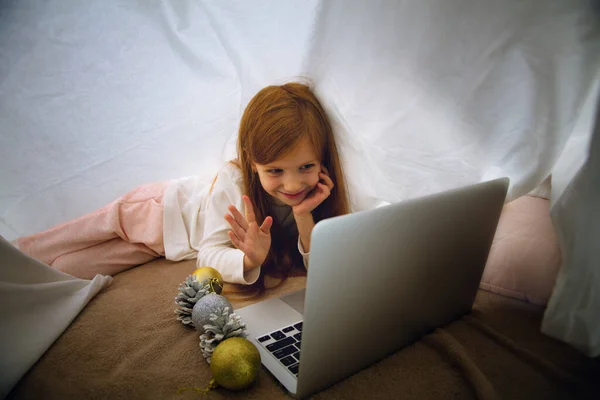 Happy caucasian little girl during video call or messaging with Santa using laptop and home devices, looks delighted and happy