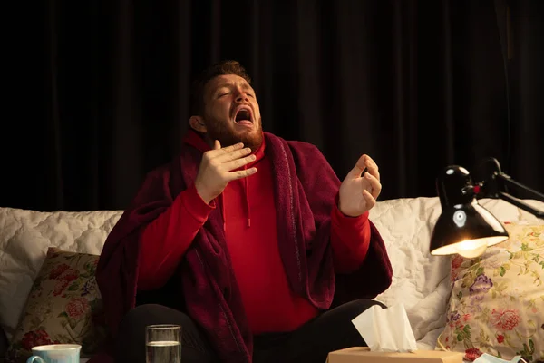 Homem envolto em xadrez parece doente, doente, espirrando e tosse sentado em casa dentro de casa — Fotografia de Stock