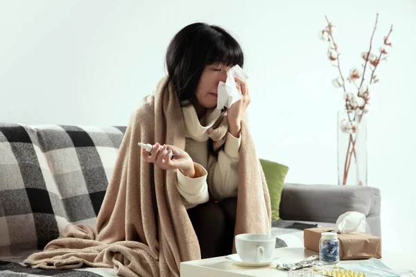Mujer envuelta en una tela a cuadros se ve enferma, enferma, estornudando y tosiendo sentada en casa dentro — Foto de Stock
