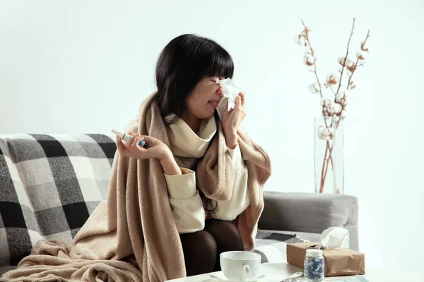 Woman wrapped in a plaid looks sick, ill, sneezing and coughing sitting at home indoors