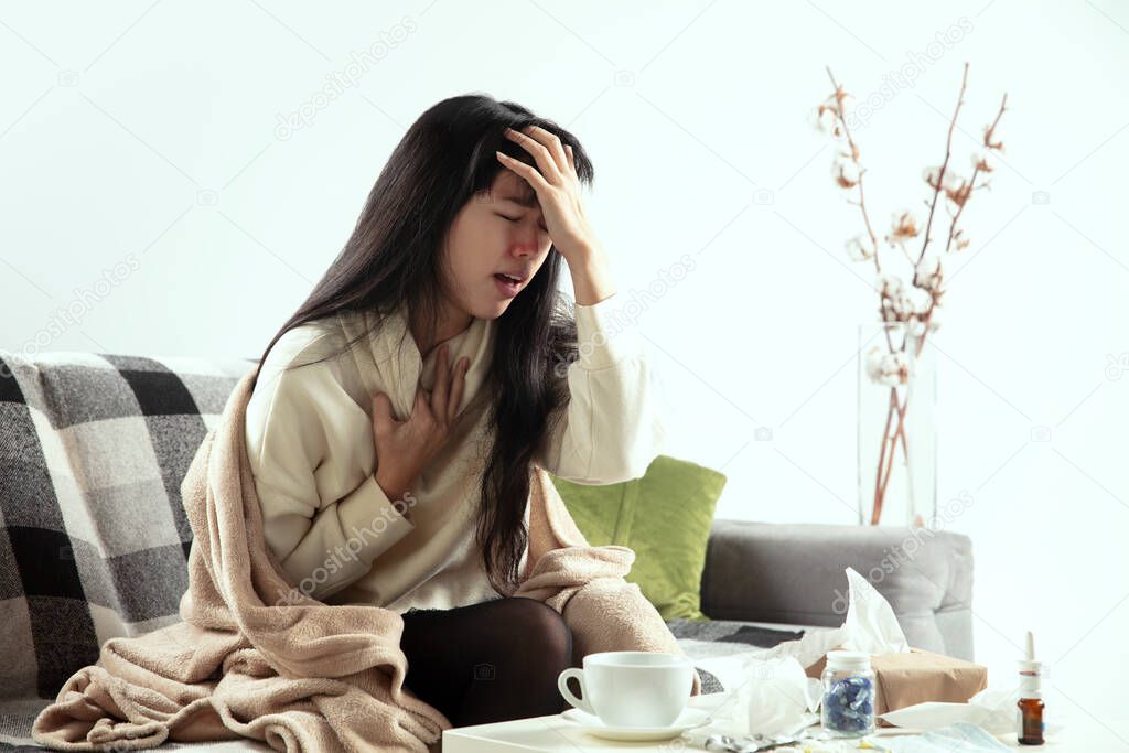 Woman wrapped in a plaid looks sick, ill, sneezing and coughing sitting at home indoors