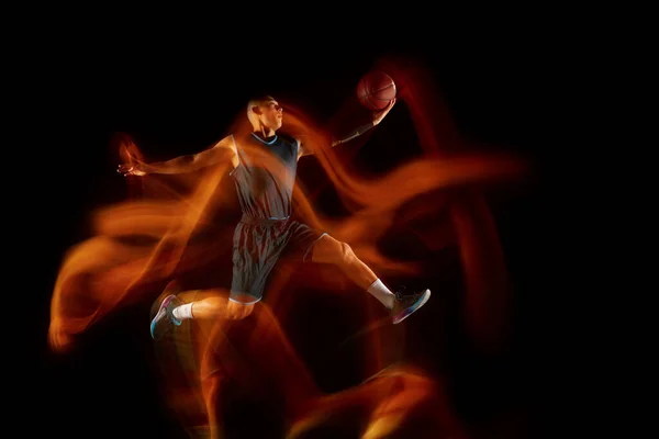 Jonge Oost-Aziatische basketbalspeler in actie en spring in gemengd licht over donkere studio-achtergrond. Begrip sport, beweging, energie en dynamische, gezonde levensstijl. — Stockfoto