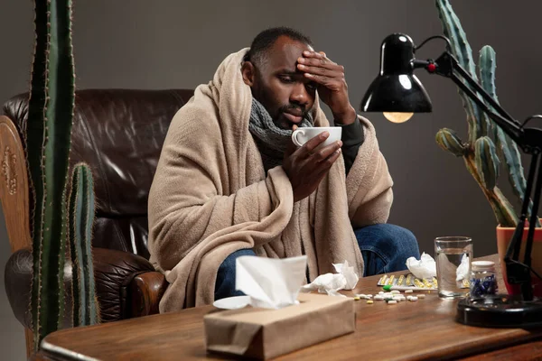 Ung afroamerikansk man insvept i en rutig ser sjuk, sjuk, har fruktansvärd huvudvärk sitter hemma. — Stockfoto
