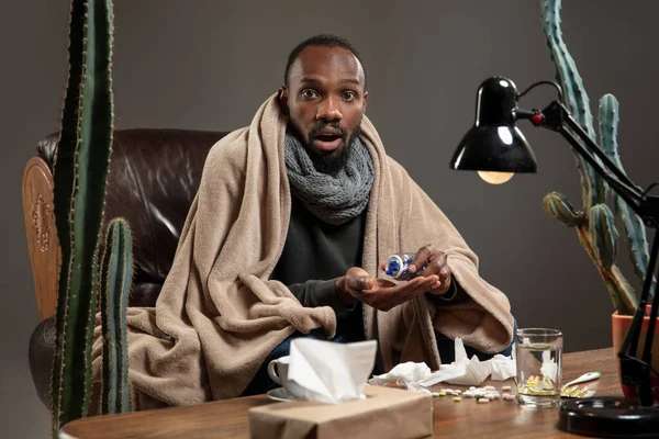 Young African-american man wrapped in a plaid looks sick, ill sitting at home indoors.