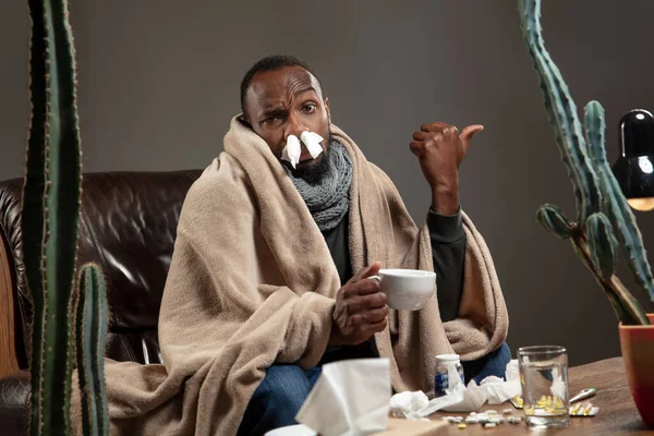 Young African-american man wrapped in a plaid looks sick, ill sitting at home indoors.