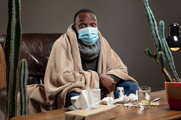Young man wrapped in a plaid looks sick, ill, sneezing and coughing sitting at home indoors in face mask.