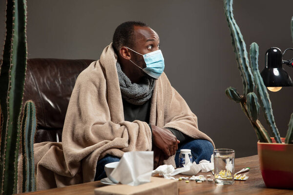 Young man wrapped in a plaid looks sick, ill, sneezing and coughing sitting at home indoors in face mask.