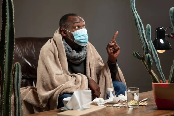 Young man wrapped in a plaid looks sick, ill, sneezing and coughing sitting at home indoors in face mask.