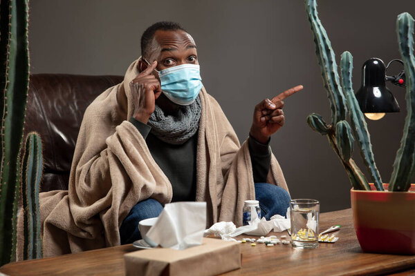 Young man wrapped in a plaid looks sick, ill, sneezing and coughing sitting at home indoors in face mask.
