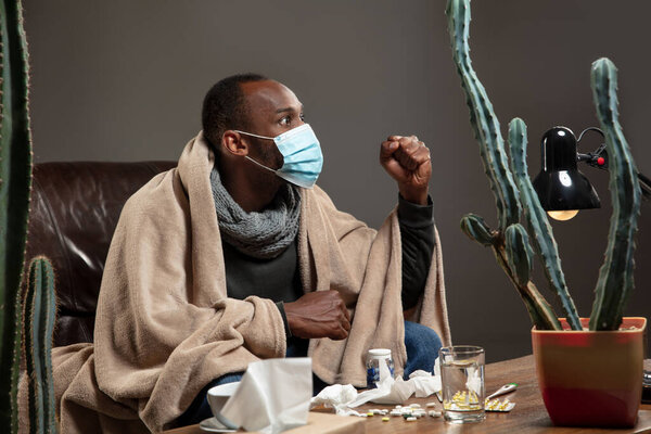 Young man wrapped in a plaid looks sick, ill, sneezing and coughing sitting at home indoors in face mask.