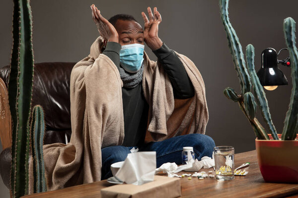 Young man wrapped in a plaid looks sick, ill, sneezing and coughing sitting at home indoors in face mask.