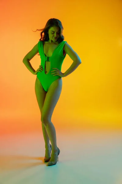 Gyönyörű csábító lány divatos zöld fürdőruha fényes gradiens sárga-narancs háttér neon fény — Stock Fotó