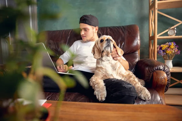 Joven hombre guapo sentado y trabajando en casa con su lindo perro. Acogedor lugar de trabajo de oficina, trabajo remoto, concepto de aprendizaje en línea. — Foto de Stock