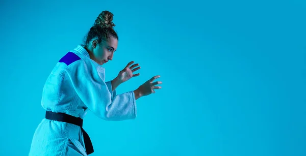 Professional female judoist training isolated on blue studio background in neon light. Healthy lifestyle, sport concept.