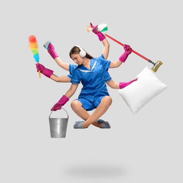 Young woman housemaid in headphones and blue uniform multitask like shiva isolated on gray background. — Stock Photo, Image