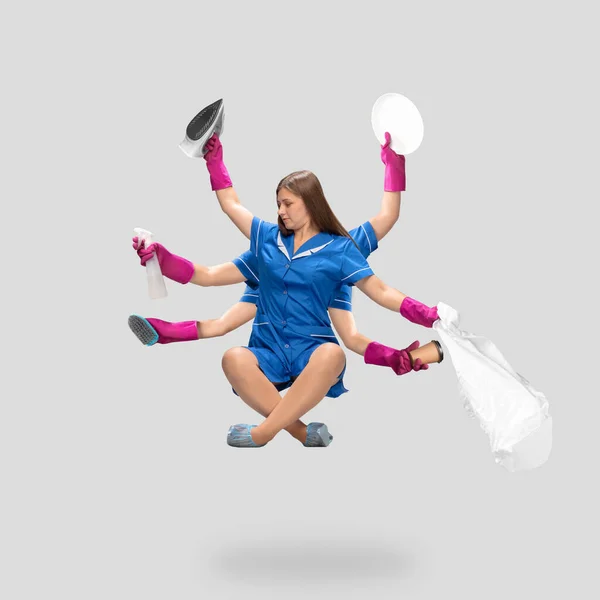 Jonge vrouw huishoudster, schoonmaakster in blauw uniform multitasken zoals shiva geïsoleerd op grijze achtergrond. — Stockfoto