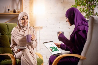 Ders sırasında evde bulunan mutlu iki Müslüman kadın, cihazlarla, online veya evde eğitim görüyorlar.