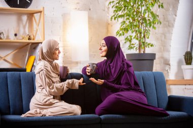 Mutlu iki Müslüman kadın evde konuşuyor, gülümsüyor, eğleniyor. Arkadaşlık, modern teknoloji, duygu kavramı.
