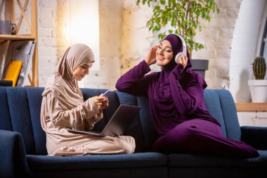 Mutlu iki Müslüman kadın evde konuşuyor, gülümsüyor, eğleniyor. Arkadaşlık, modern teknoloji, duygu kavramı.