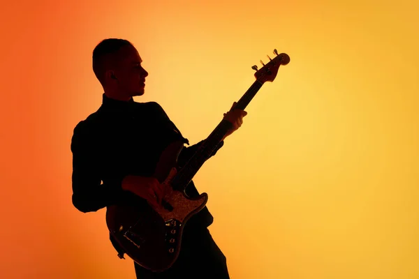 Silhueta de jovem guitarrista masculino caucasiano isolado em fundo estúdio gradiente laranja em luz de néon — Fotografia de Stock