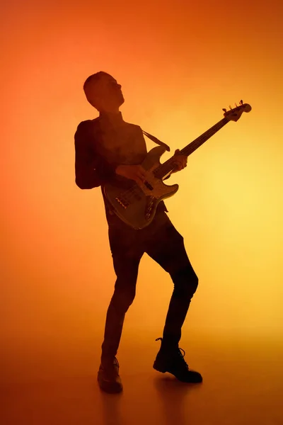 Silhouet van jonge Kaukasische mannelijke gitarist geïsoleerd op oranje gradiënt studio achtergrond in neon licht — Stockfoto
